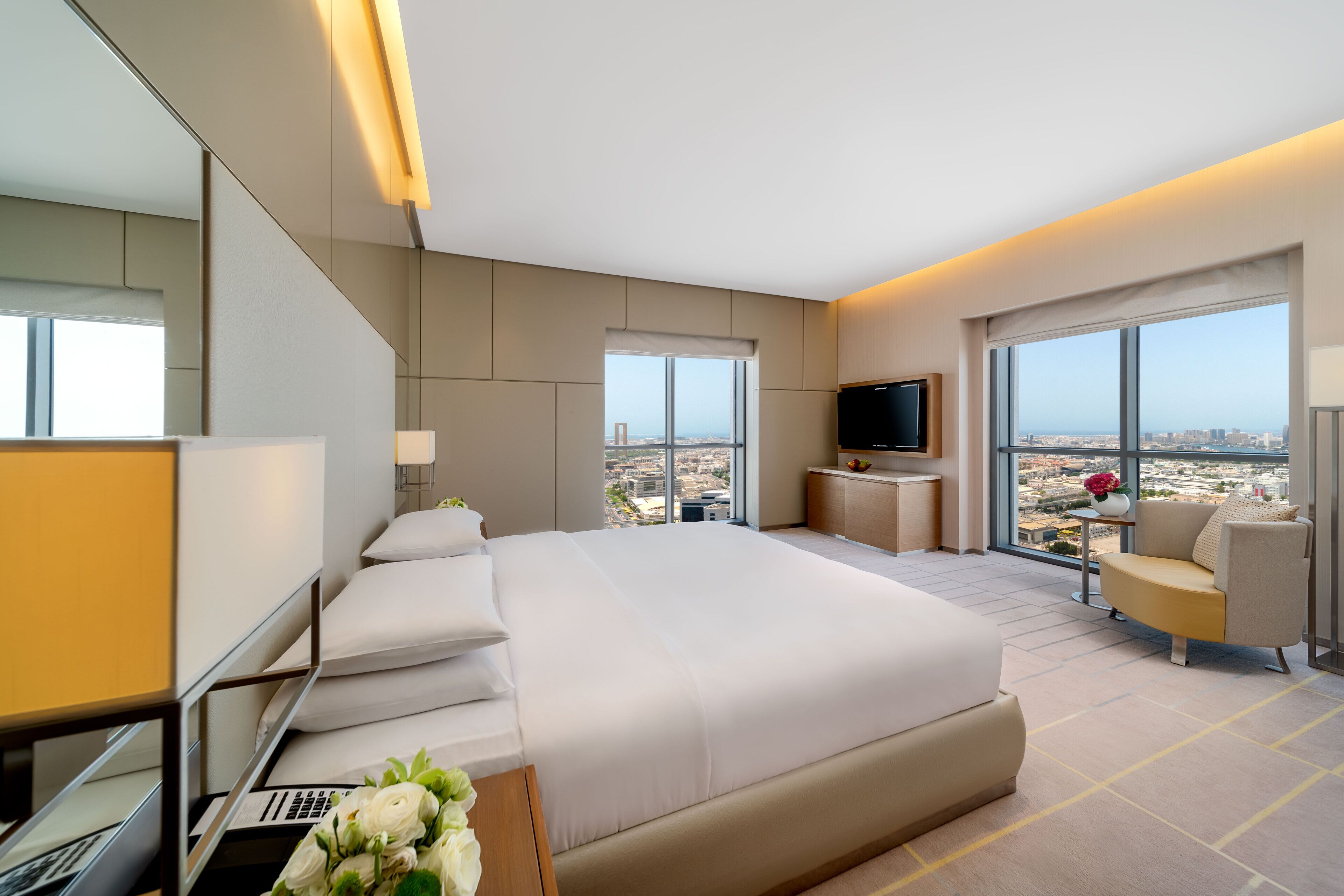 A modern hotel room with a large bed, a TV, a desk with a laptop, and a view of the city.