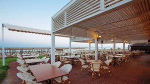 Uma área de refeições ao ar livre com várias mesas e cadeiras, um deck de madeira e vista para o mar.