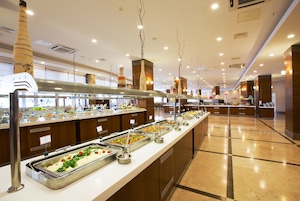 Uma área de buffet com diversas estações de comida, incluindo saladas e pratos quentes, em ambiente interno bem iluminado com detalhes em madeira e decoração moderna.