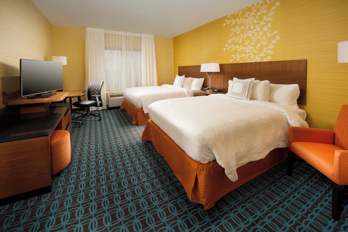 Habitación de hotel con dos camas, un escritorio con televisor, una silla y una alfombra estampada.