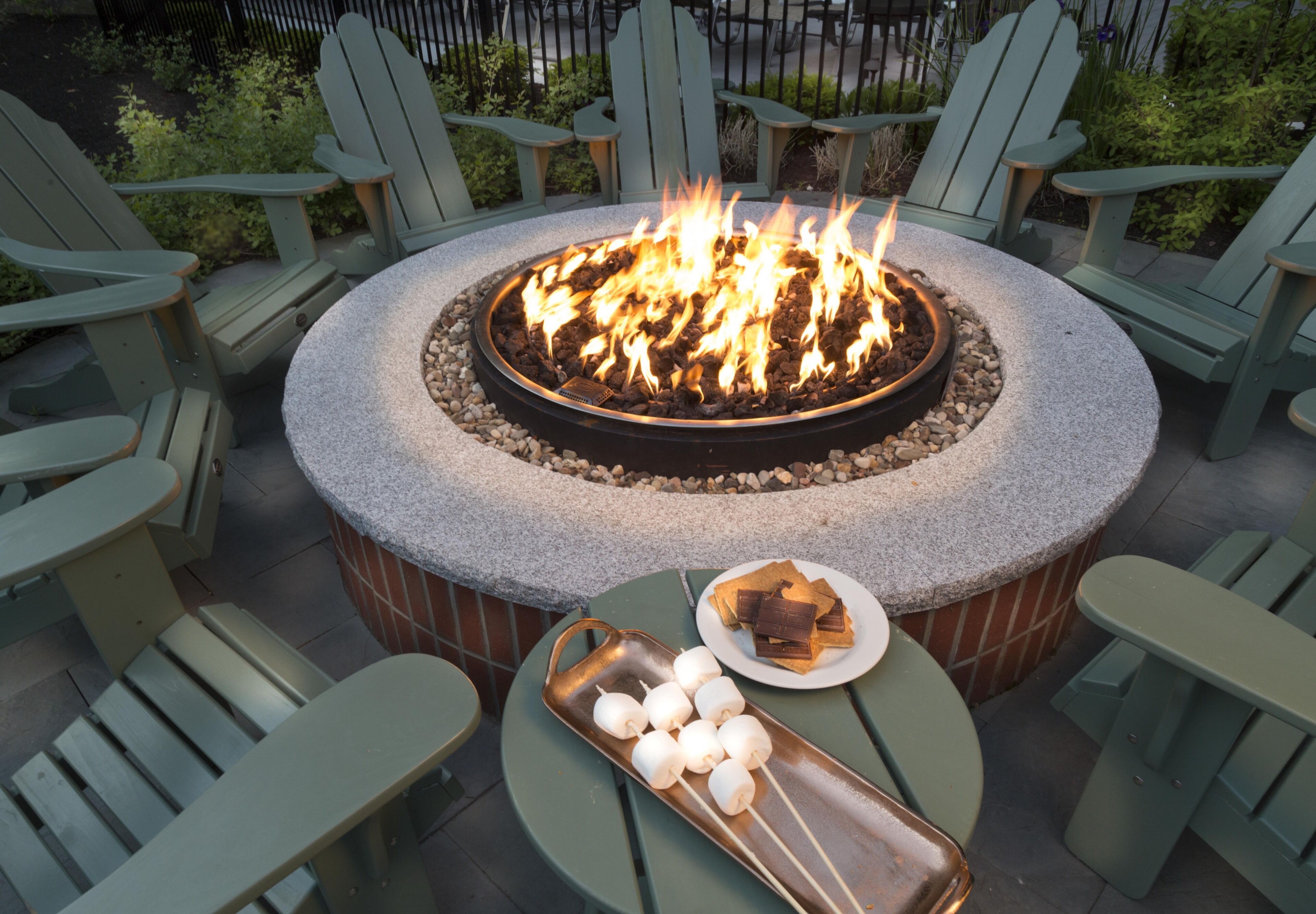 A circular outdoor seating area with a fire pit, surrounded by Adirondack chairs, and a table with a plate of chocolate and marshmallows.