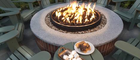 A circular outdoor seating area with a fire pit, surrounded by Adirondack chairs, and a table with a plate of chocolate and marshmallows.