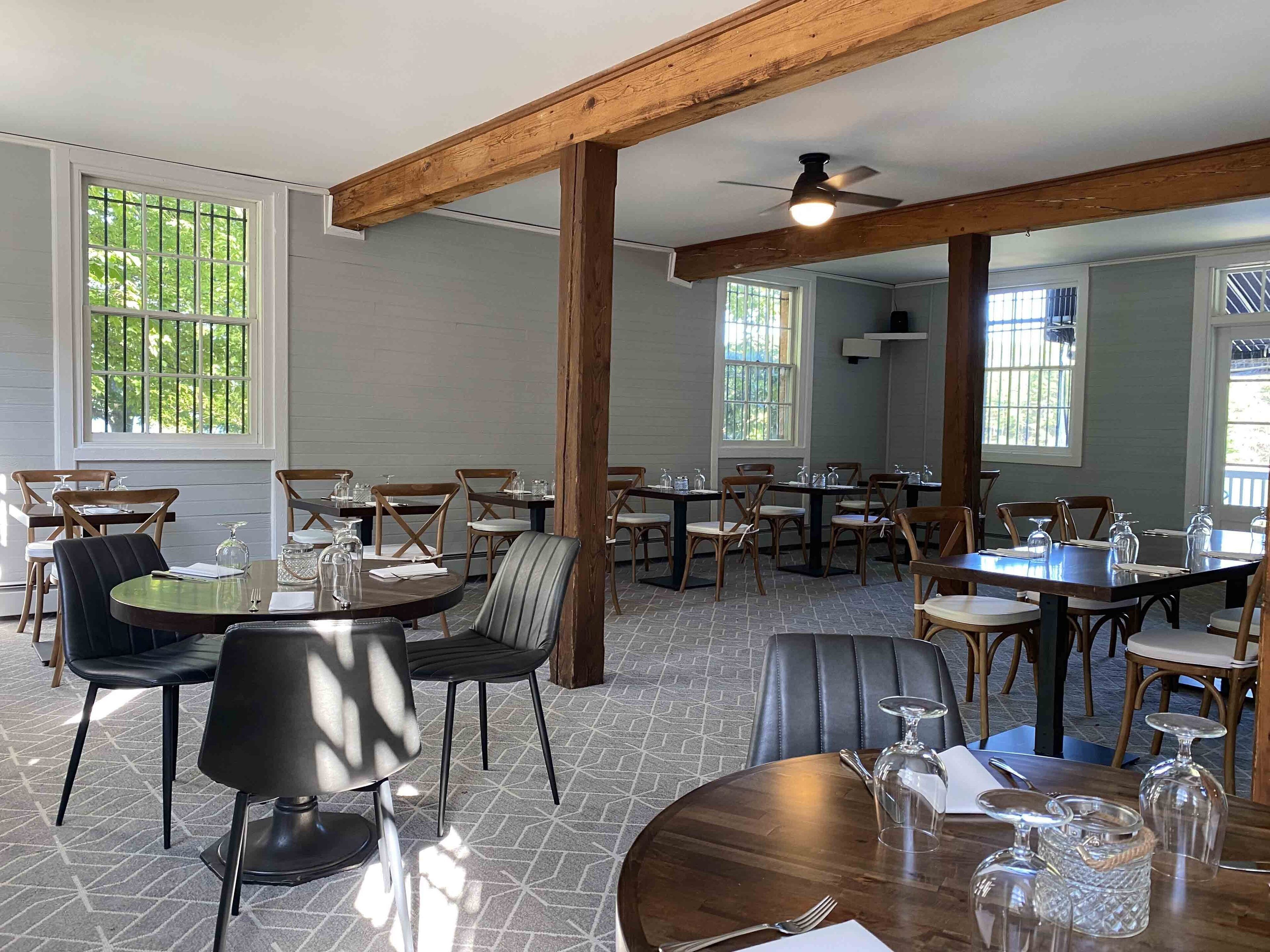 A restaurant with wooden beams, large windows, and a variety of seating arrangements including round tables and chairs.