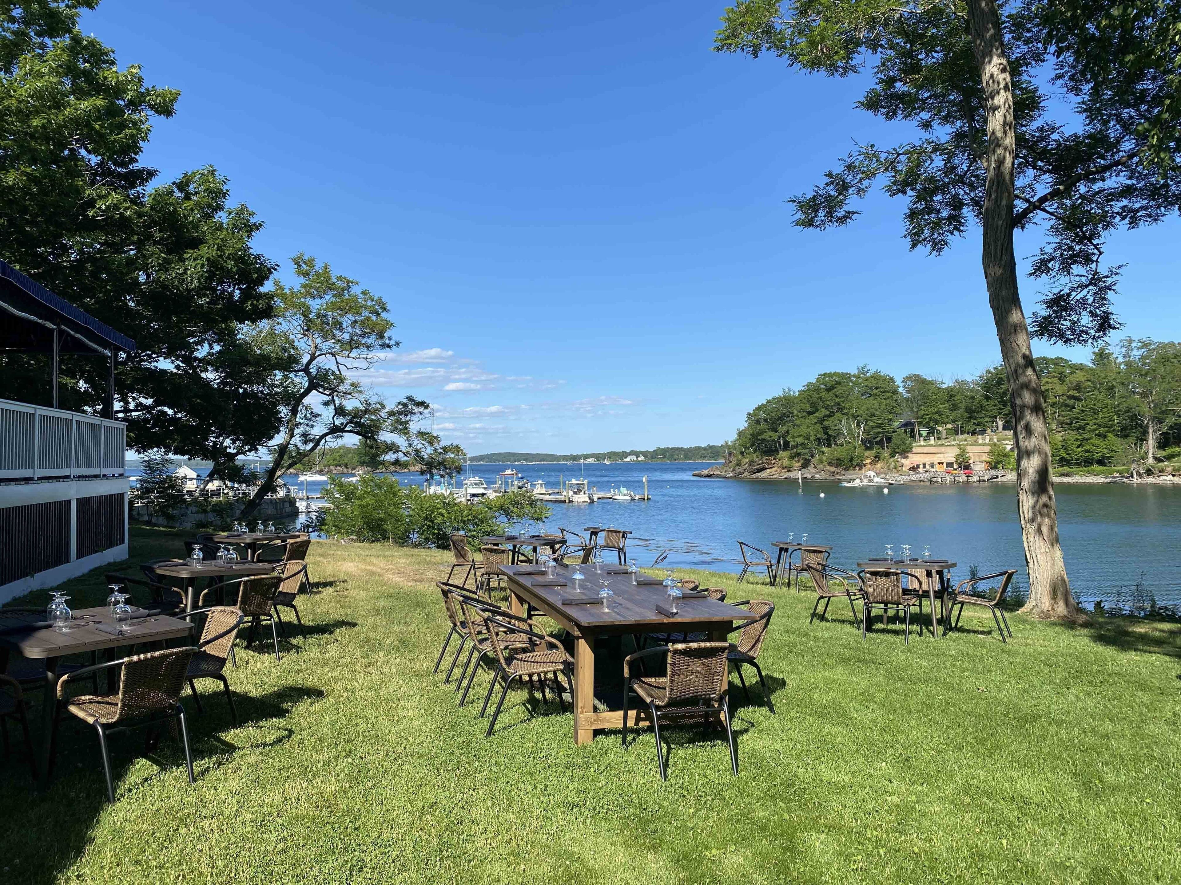 An outdoor dining area with tables and chairs, overlooking a body of water with boats and a dock.