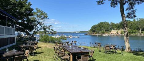 An outdoor dining area with tables and chairs, overlooking a body of water with boats and a dock.