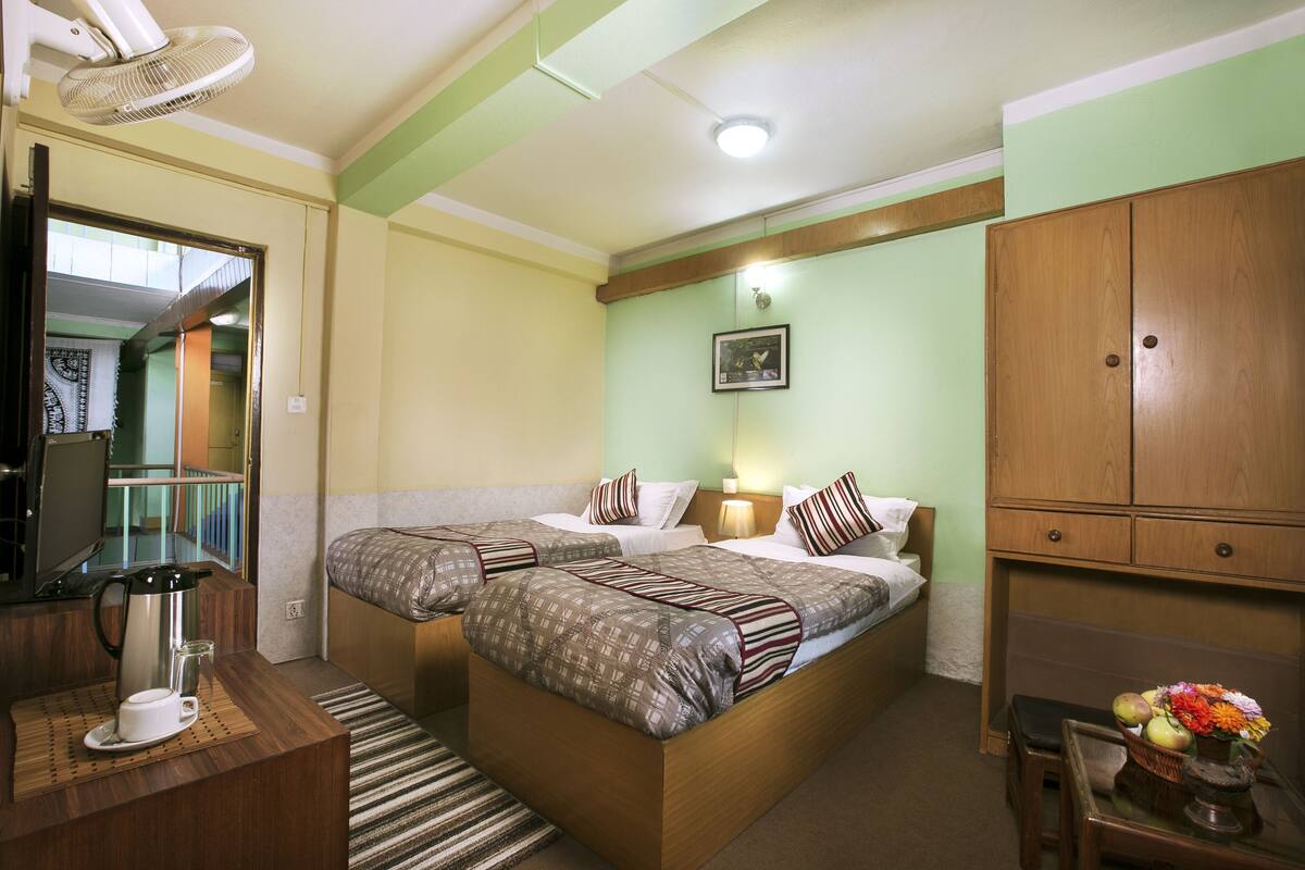 A hotel room with two beds, a desk with a coffee maker, a mirror, a wardrobe, and a small table with flowers.