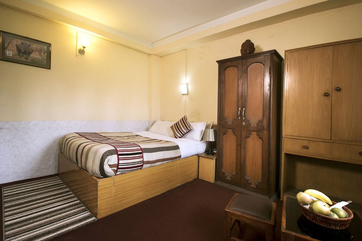 A hotel room with a bed, a wooden wardrobe, a nightstand, a lamp, a small table, and a basket of fruit.