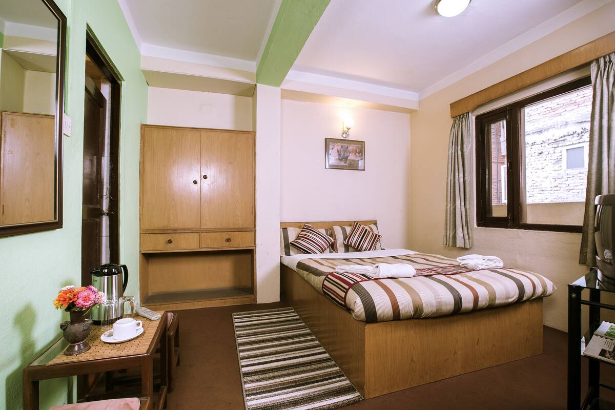 A hotel room with a bed, a wooden wardrobe, a desk with a coffee maker, a small table with flowers, and a window with curtains.