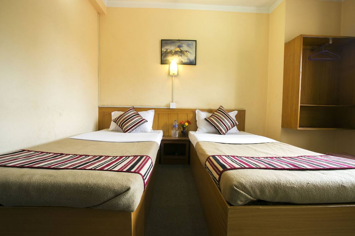 A hotel room with two single beds, a nightstand with a lamp, a painting on the wall, and a wooden wardrobe.