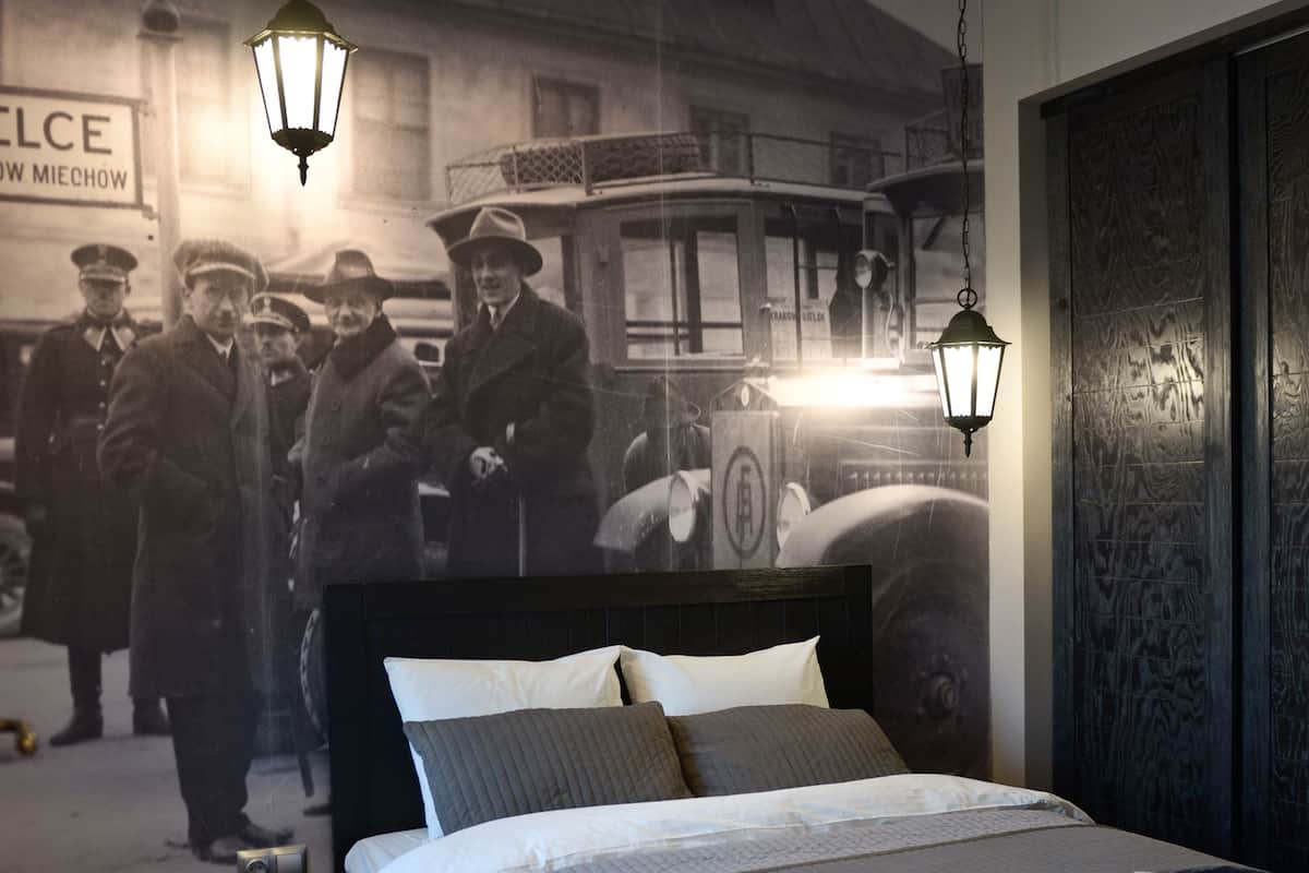 Um quarto com uma cama, uma mesa de cabeceira, um relógio de parede e um cartaz antigo.