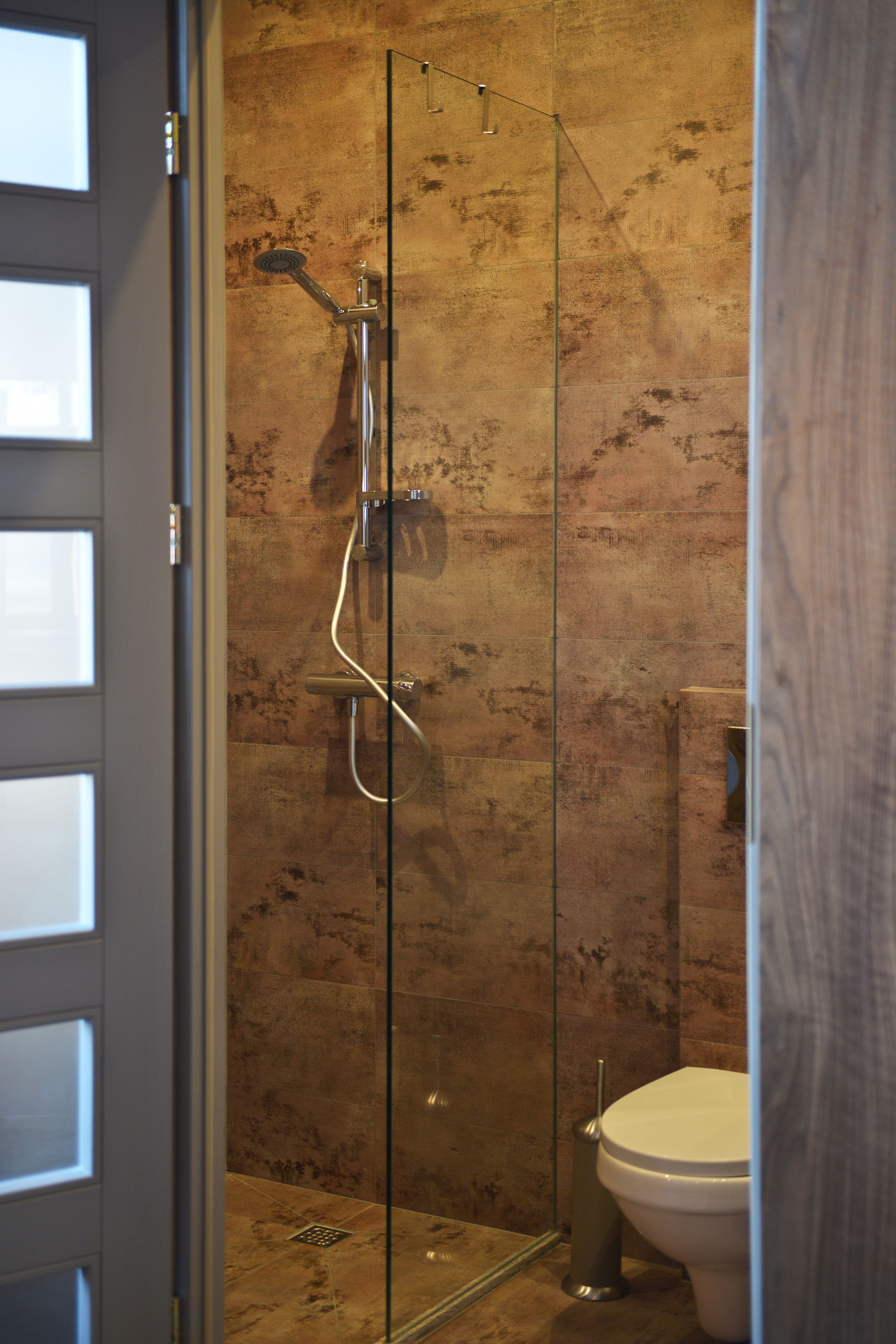 Ein Duschbereich mit Glastrennwand, einer Duschbrause und einer Bedienkonsole, neben einer Toilette in einem Badezimmer mit strukturierter Wand.