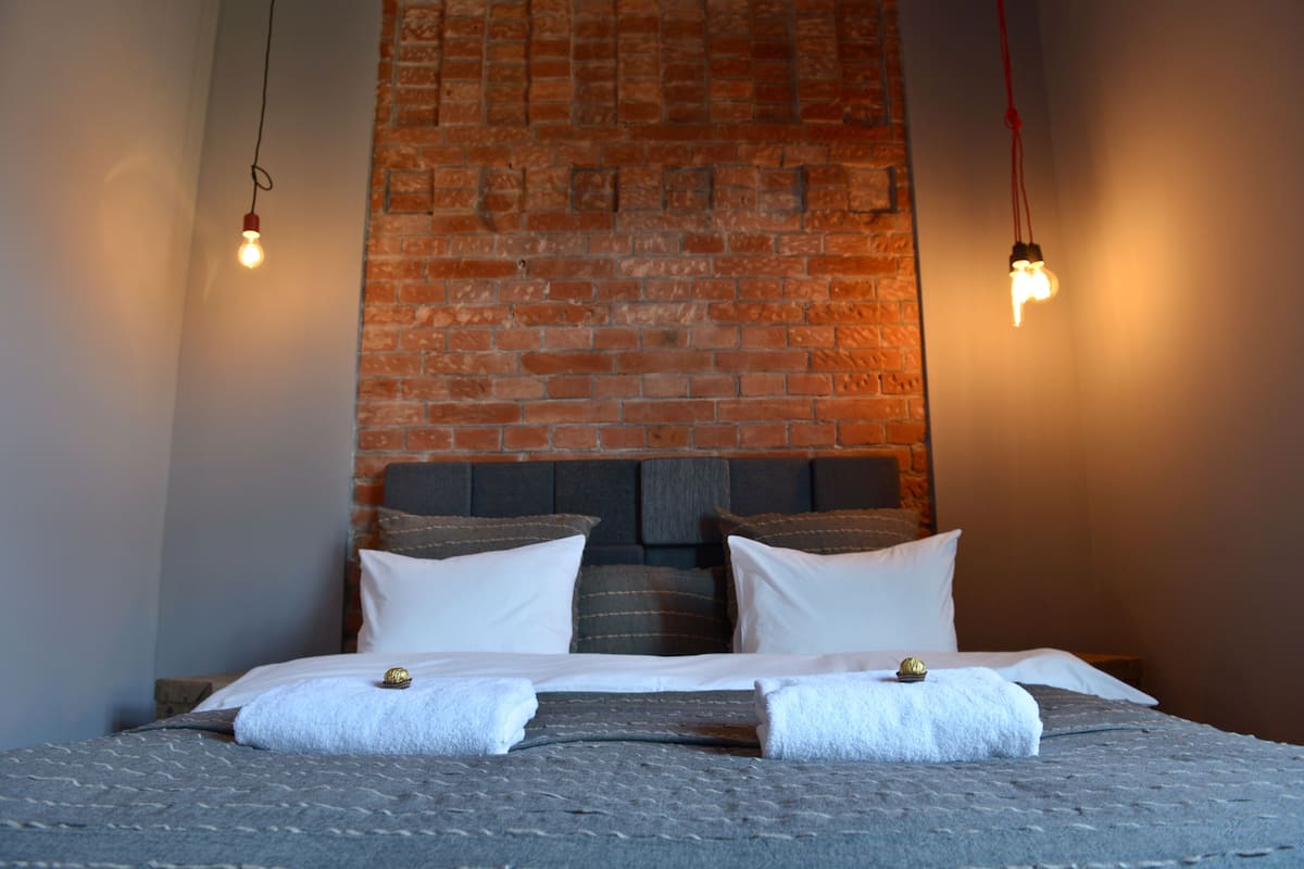 Uma cama com lençóis brancos, uma parede de tijolos ao fundo e duas lâmpadas penduradas.