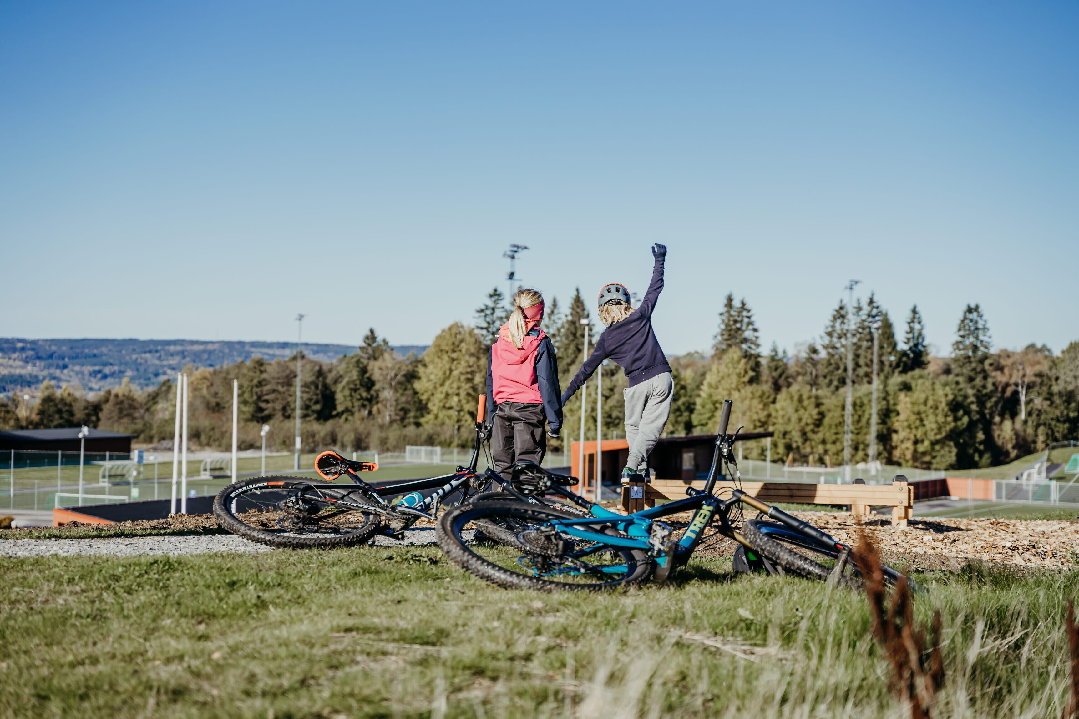 Two people standing next to a mountain bike on a grassy field with trees and a clear sky in the background.