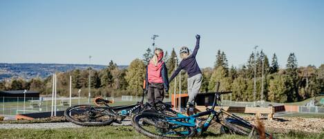 Two people standing next to a mountain bike on a grassy field with trees and a clear sky in the background.
