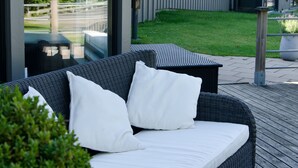 Een rieten bank met witte kussens op een terras, een glazen deur die uitkomt op een tuin, en een modern gebouw met een rood dak op de achtergrond.