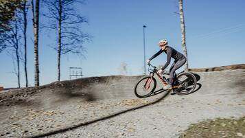A person riding a bicycle on a dirt path with trees and a clear sky in the background.