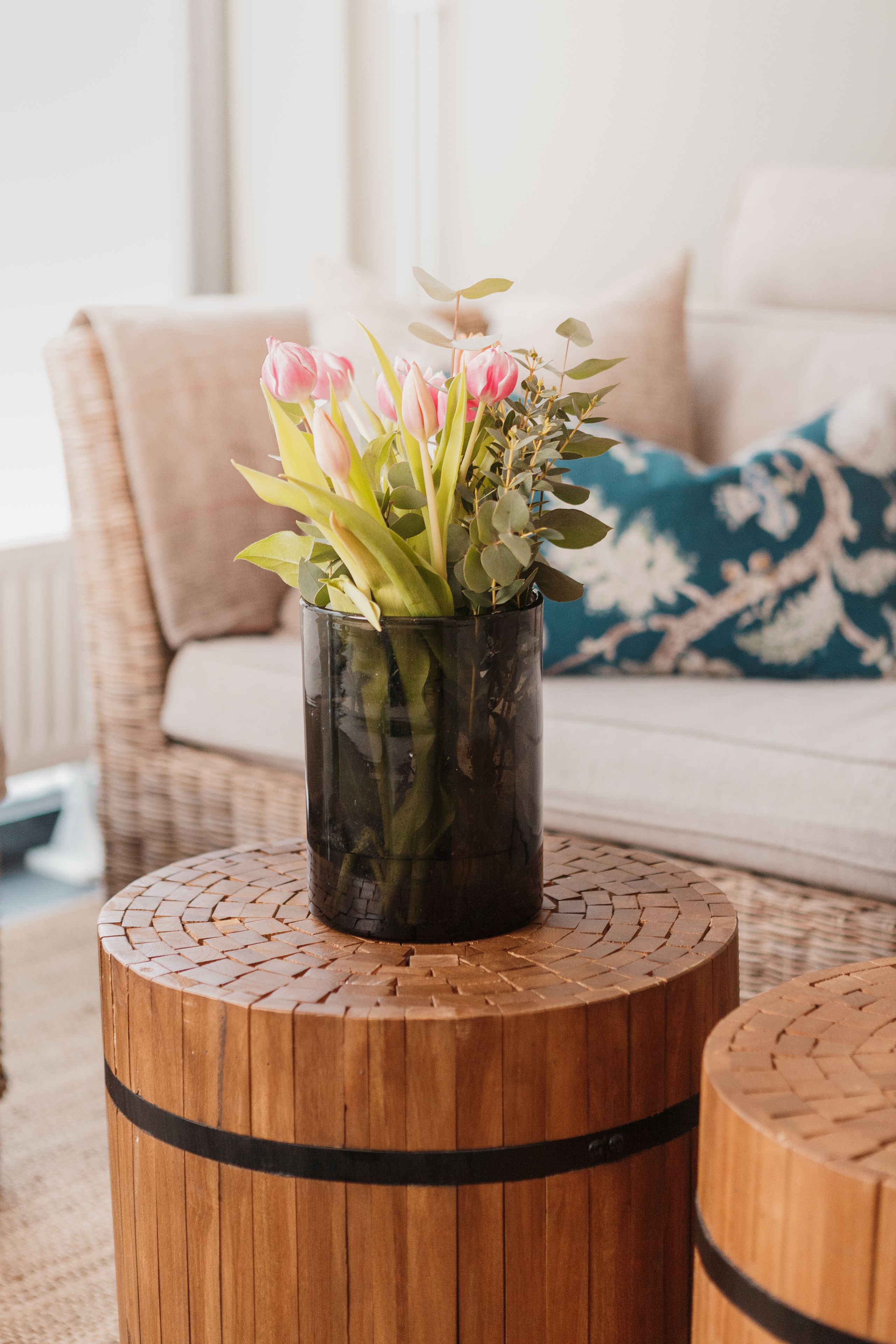 A vase with pink flowers on a wooden stool in a living room with a sofa and a patterned pillow.