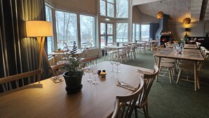 Een restaurant met houten tafels en stoelen, grote ramen en hangende lampen.