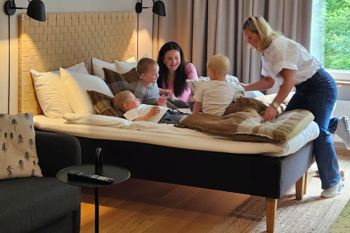 Ein Schlafzimmer mit Bett, Nachttisch, Lampe, einem Fenster mit Vorhängen und Blick ins Grüne.