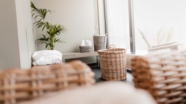 Een kamer met een groot raam, een plant, rieten manden en een opgemaakt bed.