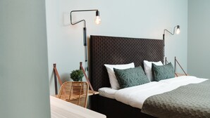 Een moderne slaapkamer met een donker gewatteerd hoofdbord, een houten nachtkastje, een rieten stoel en twee wandlampen voor het lezen.