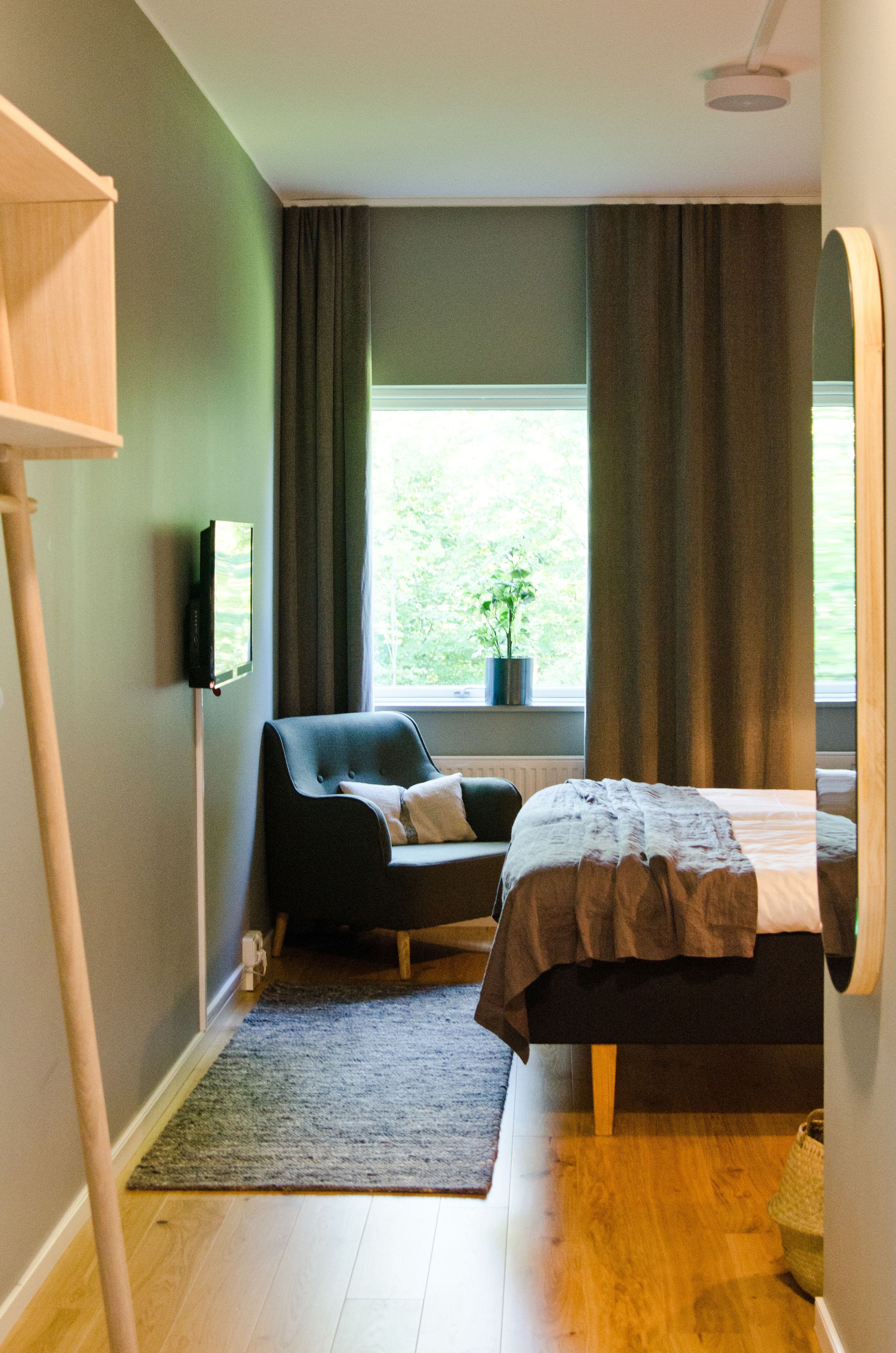 Ein Schlafzimmer mit einem Bett, einem Sessel, einem Fenster mit Vorhängen, einer Pflanze und einem flachbildfernseher an der Wand.