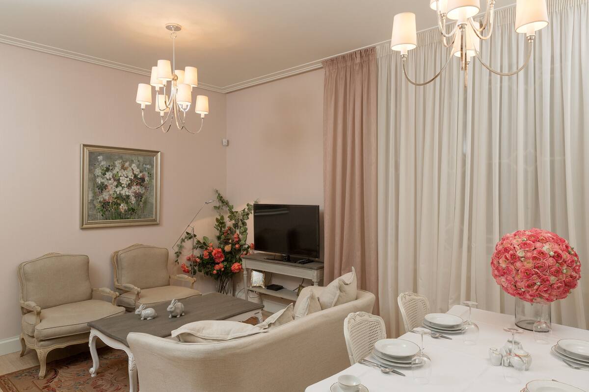 Un salon avec un téléviseur, des meubles de couleur beige, une table à manger dressée pour quatre personnes et un lustre.