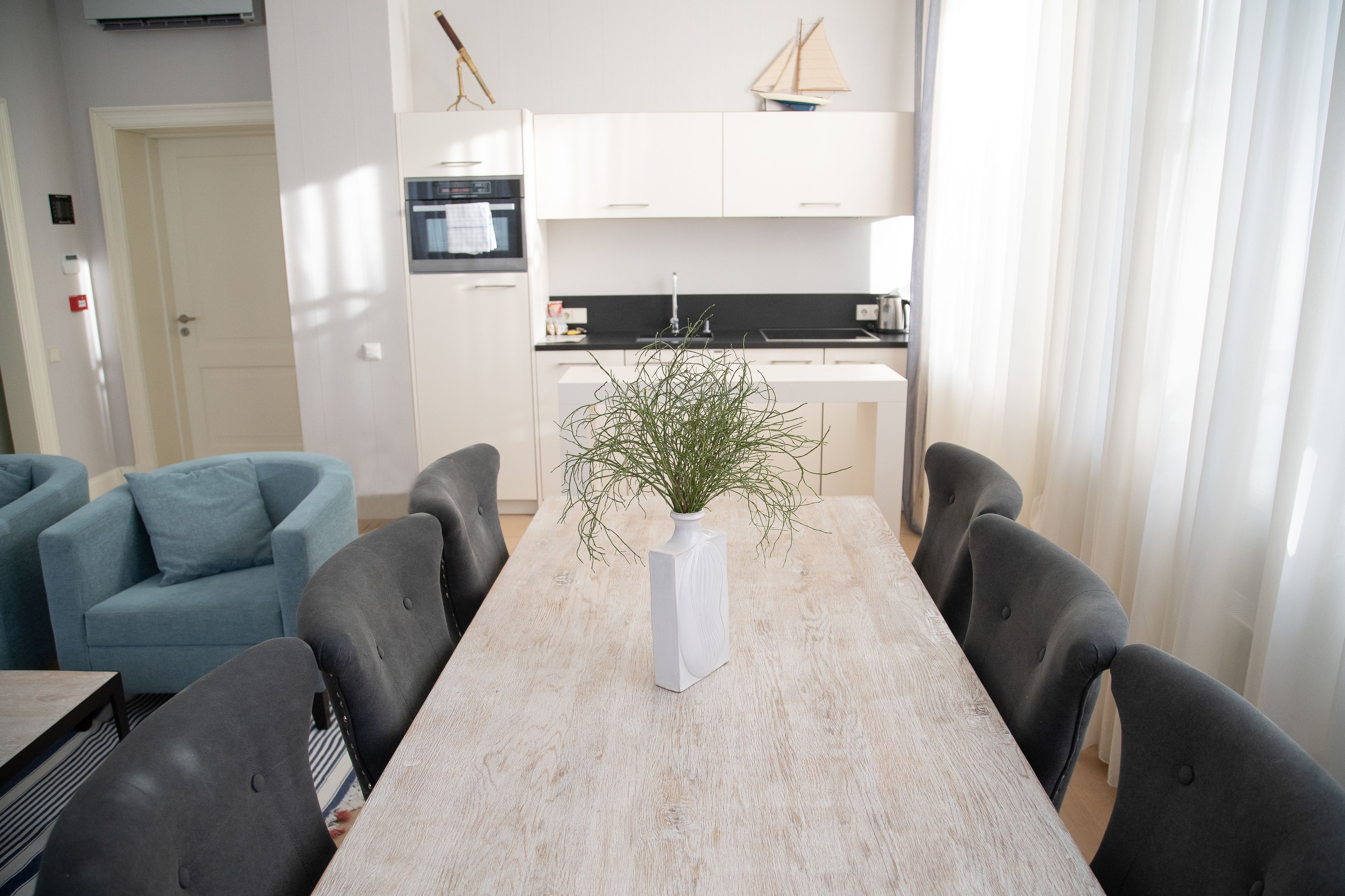 Un comedor con una mesa de madera, cuatro sillas, un jarrón blanco con follaje y una cocina al fondo.