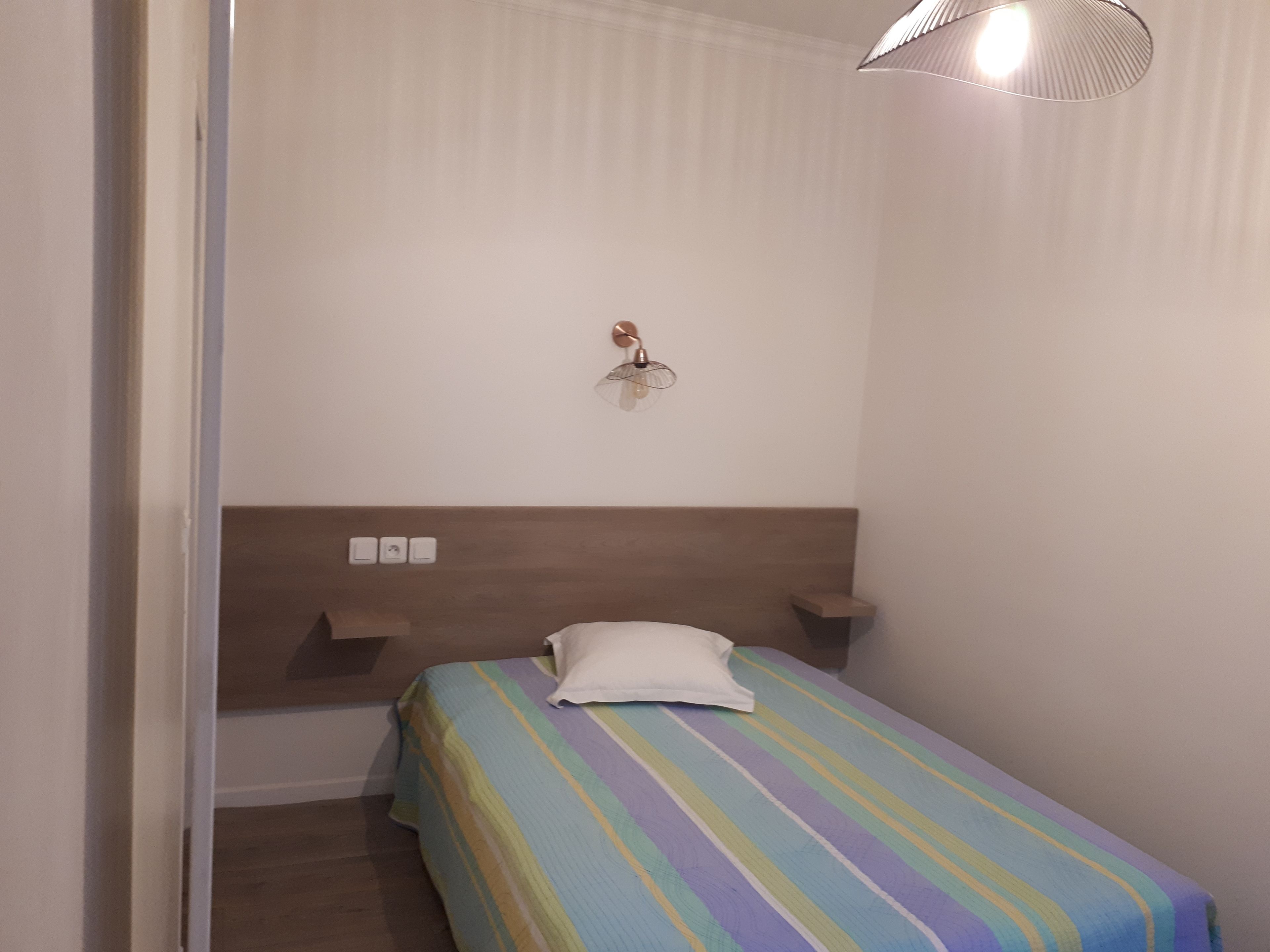 Ein kleines, ordentlich eingerichtetes Schlafzimmer mit einem Einzelbett, einem Nachttisch, einer Wandleuchte und einer Steckdose.
