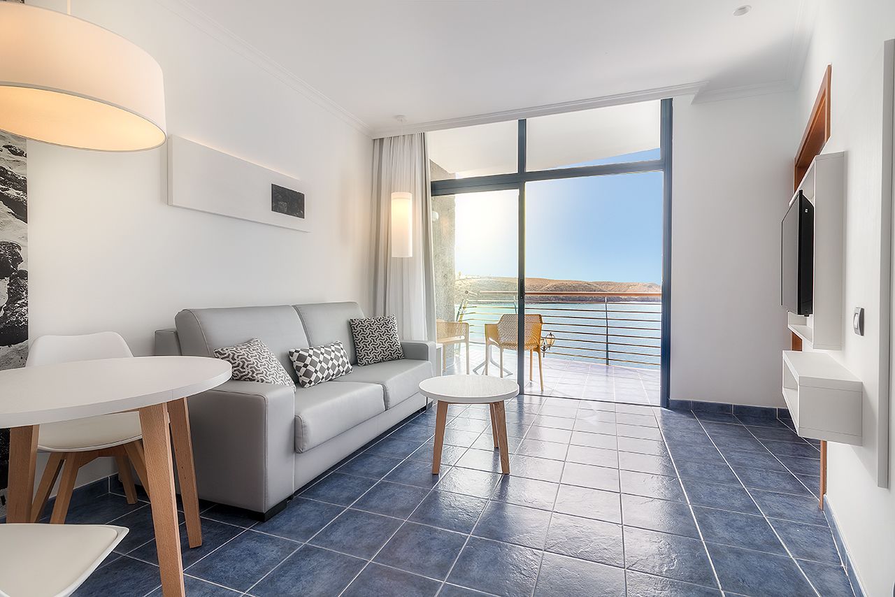Habitación de hotel moderna con sofá, mesa de comedor y balcón con vista al mar.