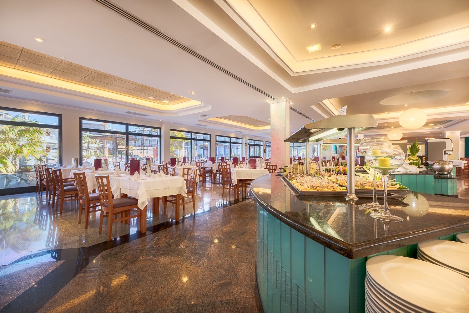 A spacious restaurant with multiple dining tables and chairs, a central buffet area, and large windows offering a view of the outdoors.