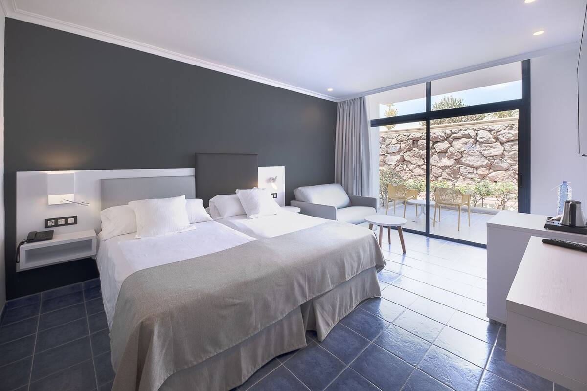 A modern hotel room with two beds, a desk, and a balcony with a stone wall view.