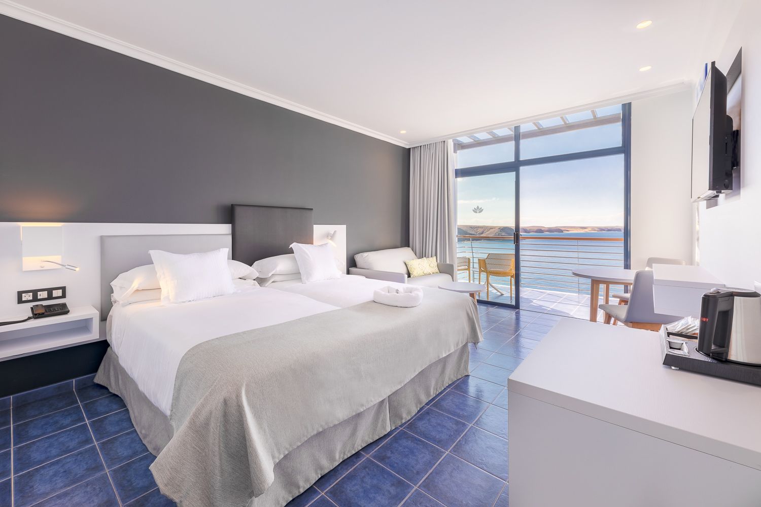 A modern hotel room with a large bed, a desk with a coffee maker, and a balcony with a view of the sea.