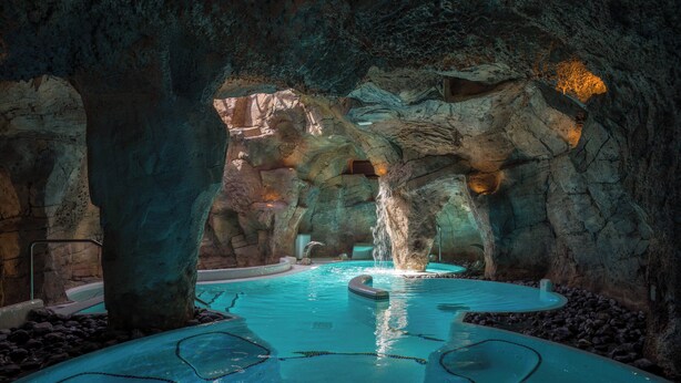 Une piscine intérieure aménagée dans une grotte naturelle, avec une fontaine centrale et plusieurs espaces de détente.