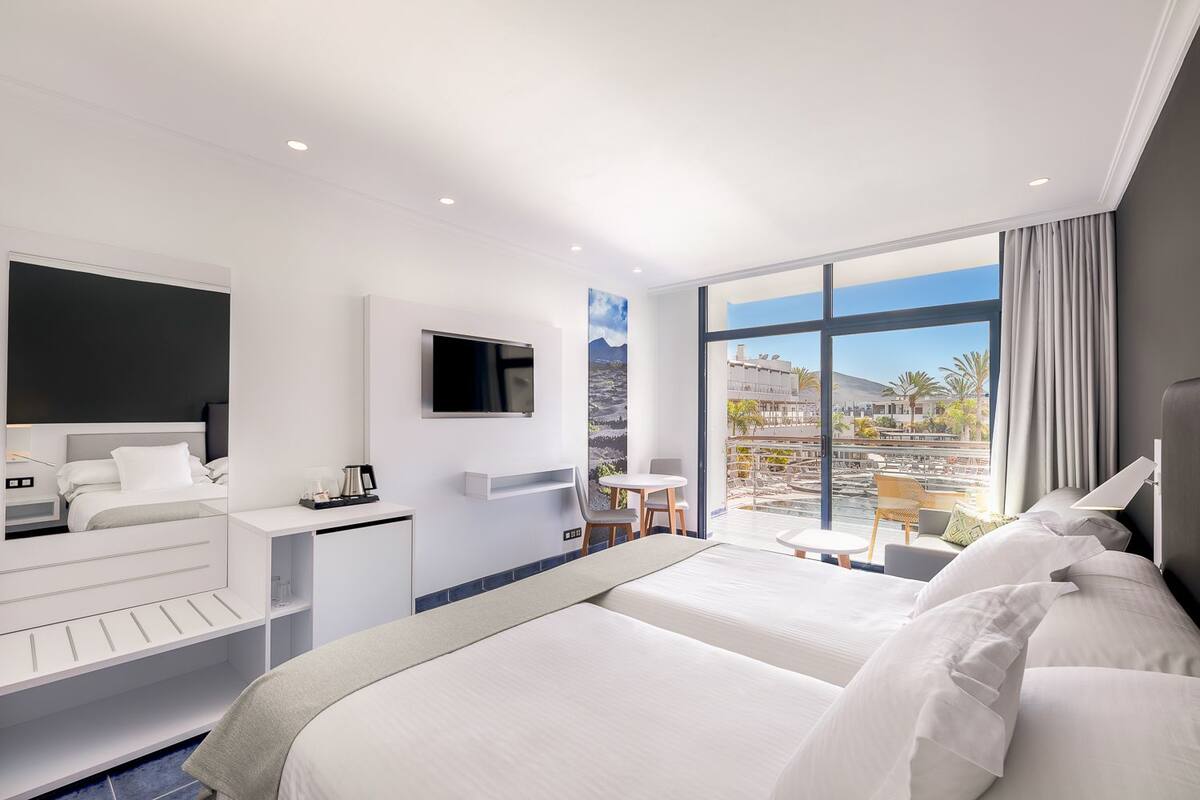A modern hotel room with a large bed, a desk, a TV, and a balcony with a view of a pool and buildings.