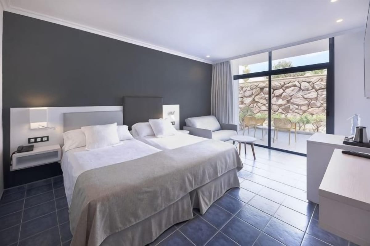 Une chambre d’hôtel moderne avec deux lits, un bureau et une grande fenêtre donnant sur un mur en pierre.