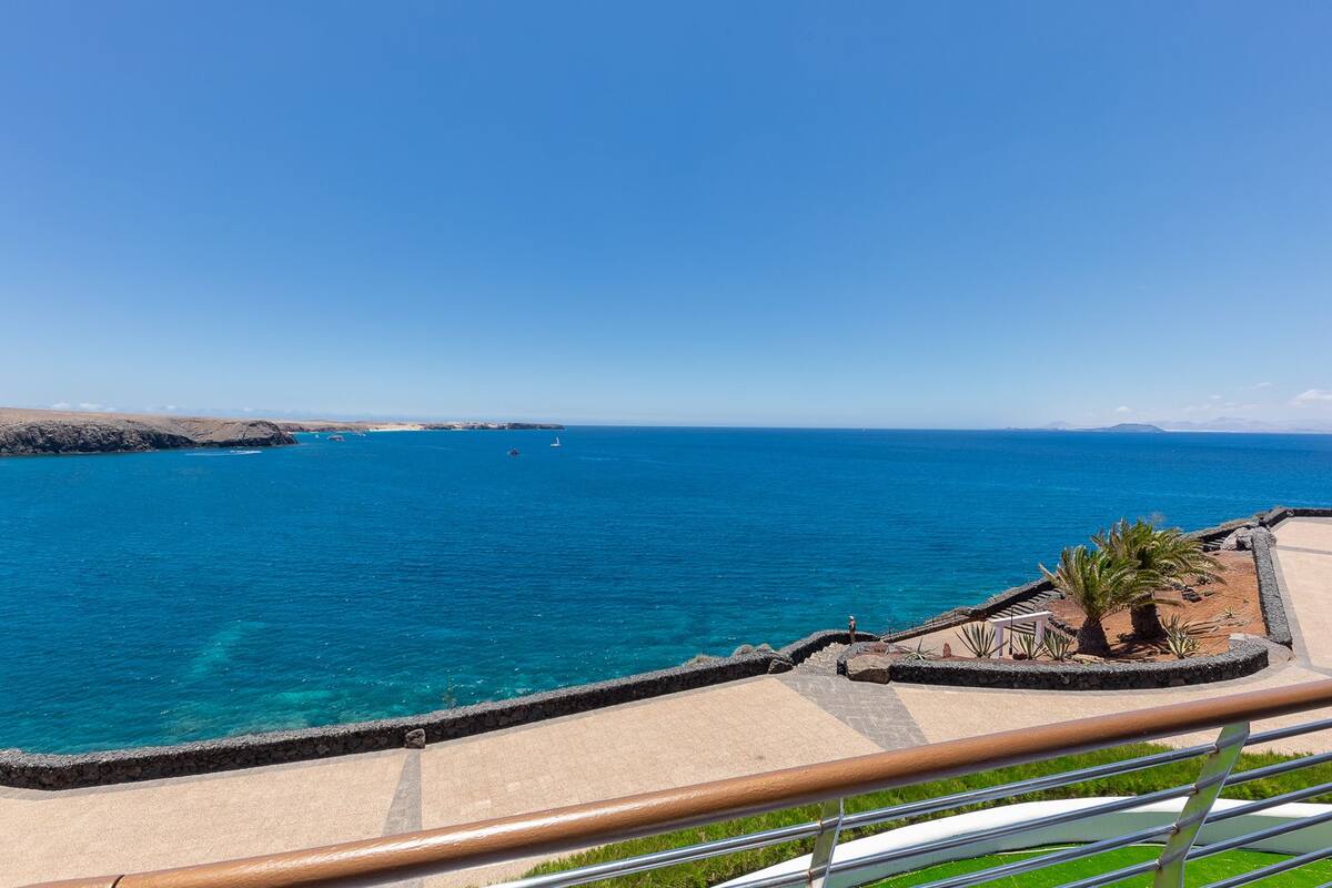 A coastal view with a clear blue sea, a sandy beach, and a stone wall.