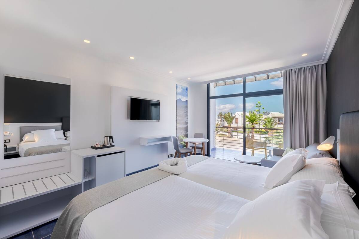 A modern hotel room with a large bed, a desk, a TV, and a balcony with a view.
