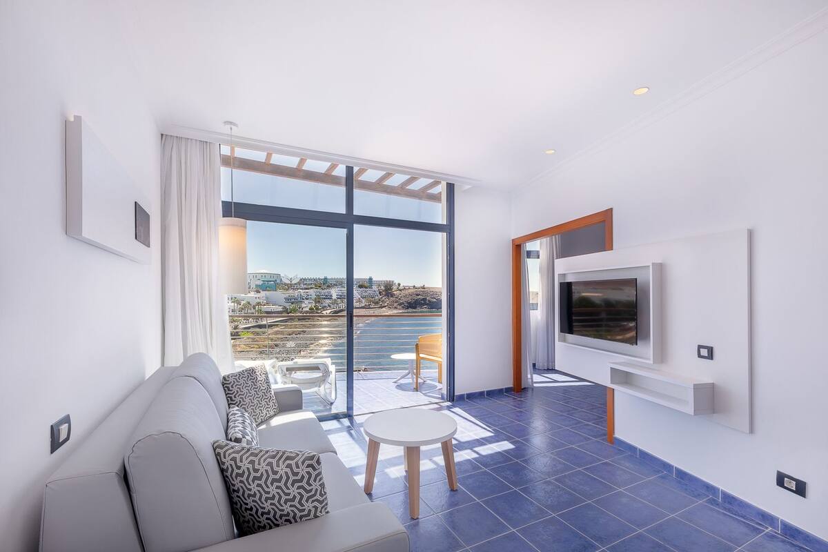 A modern living room with a sofa, a small table, and a TV mounted on the wall. There is a large window offering a view of the city and a balcony with outdoor furniture.