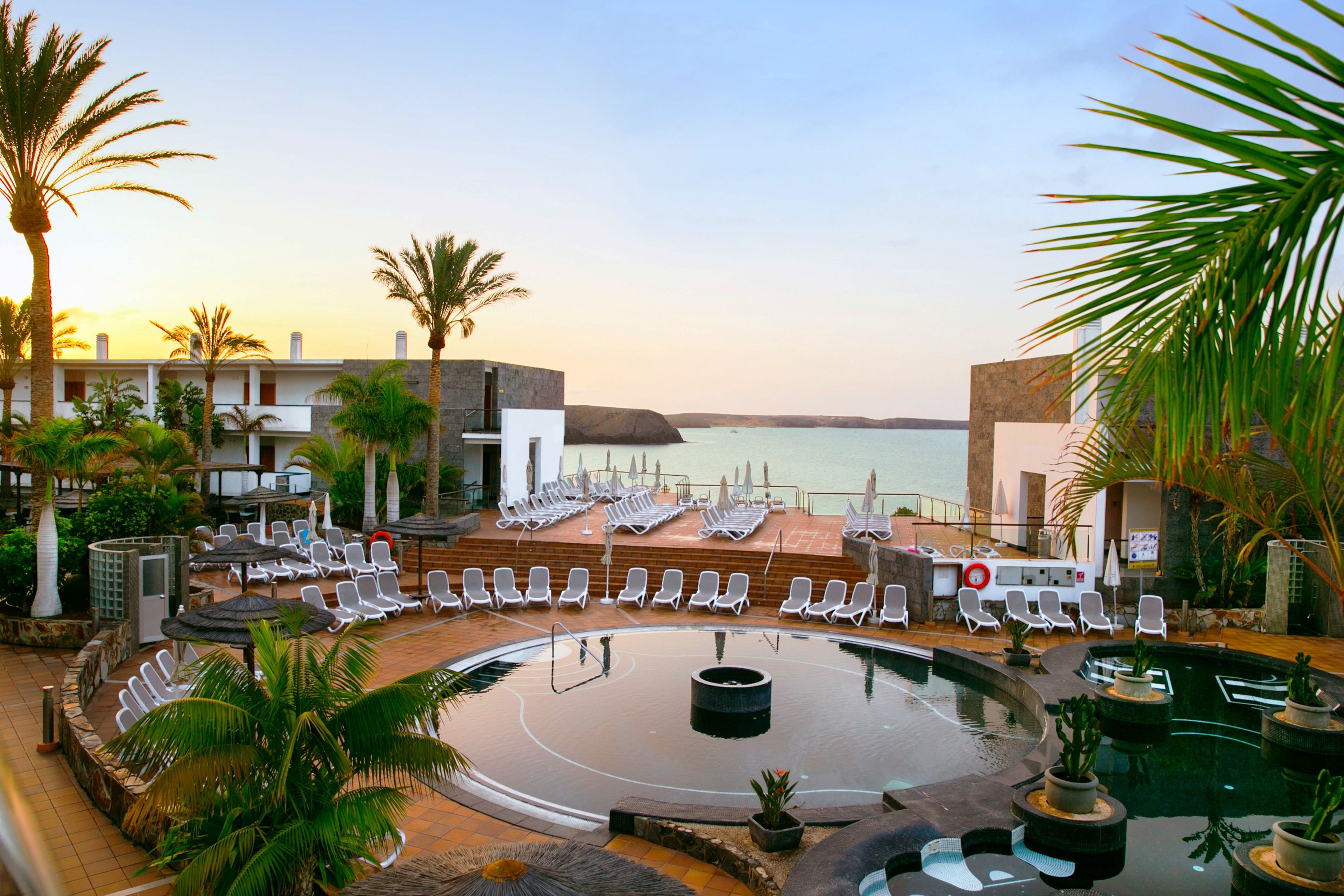 A pool area with lounge chairs and palm trees, overlooking a body of water.