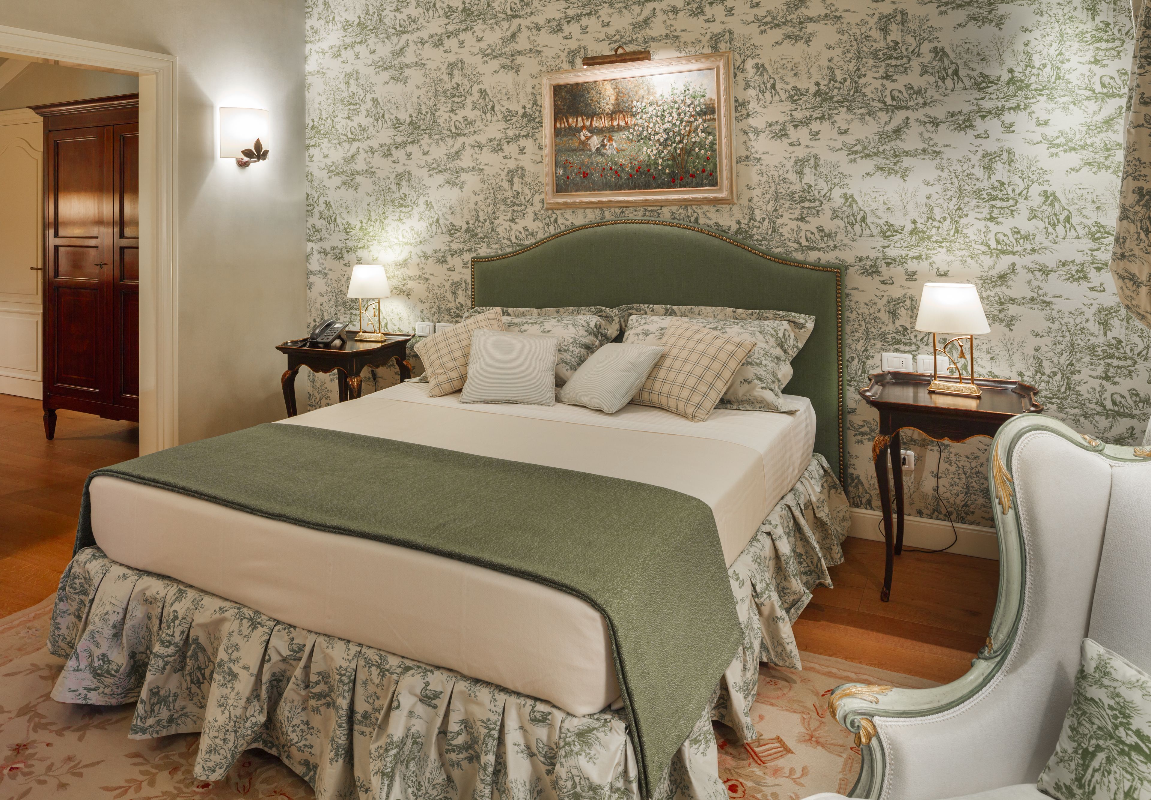 A bedroom with a large bed, floral wallpaper, a green headboard, two bedside tables with lamps, and a chair.
