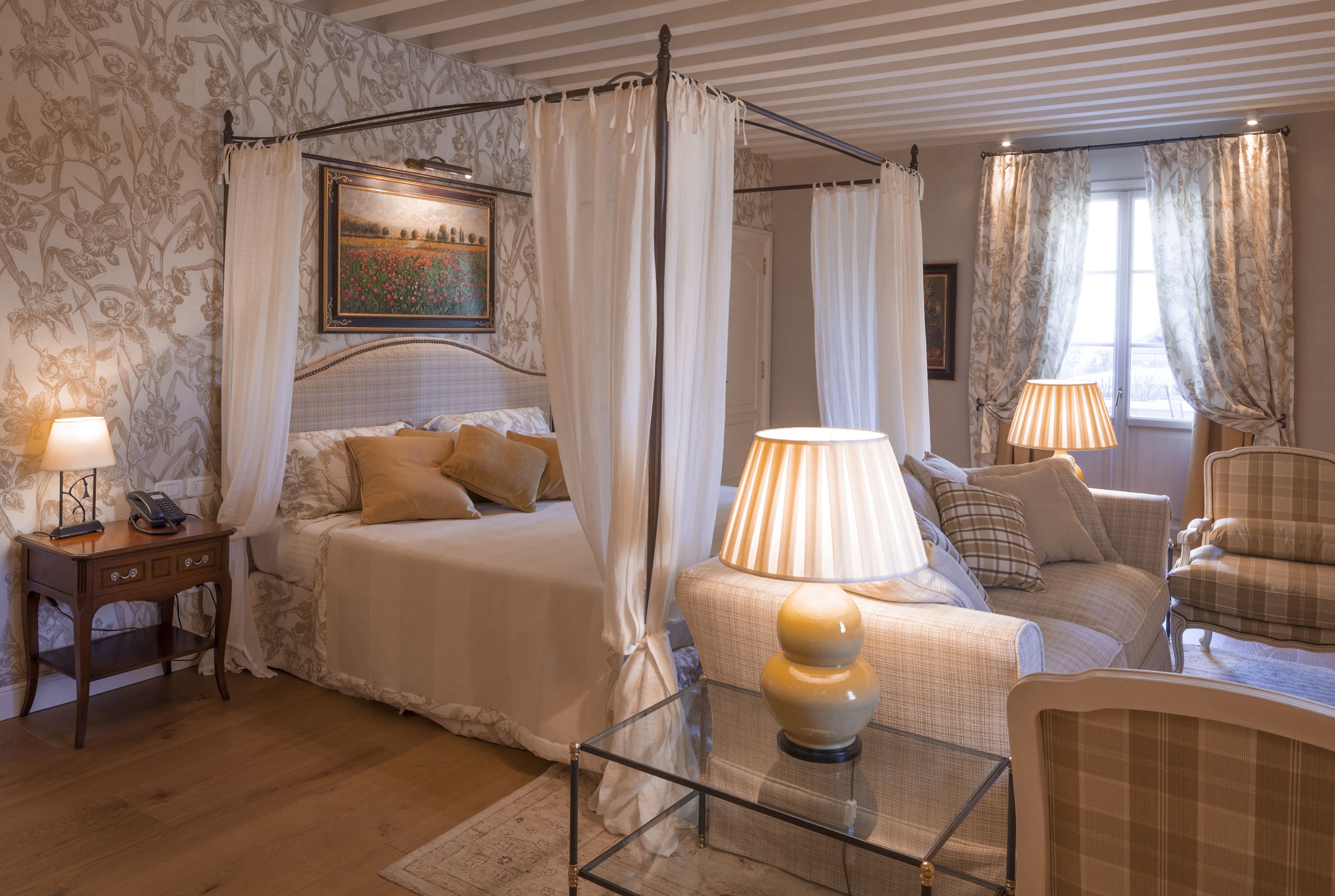 Un dormitorio con cama con dosel, una zona de estar con sofá y sillones, una mesa de centro de cristal y una mesita de noche con lámpara.