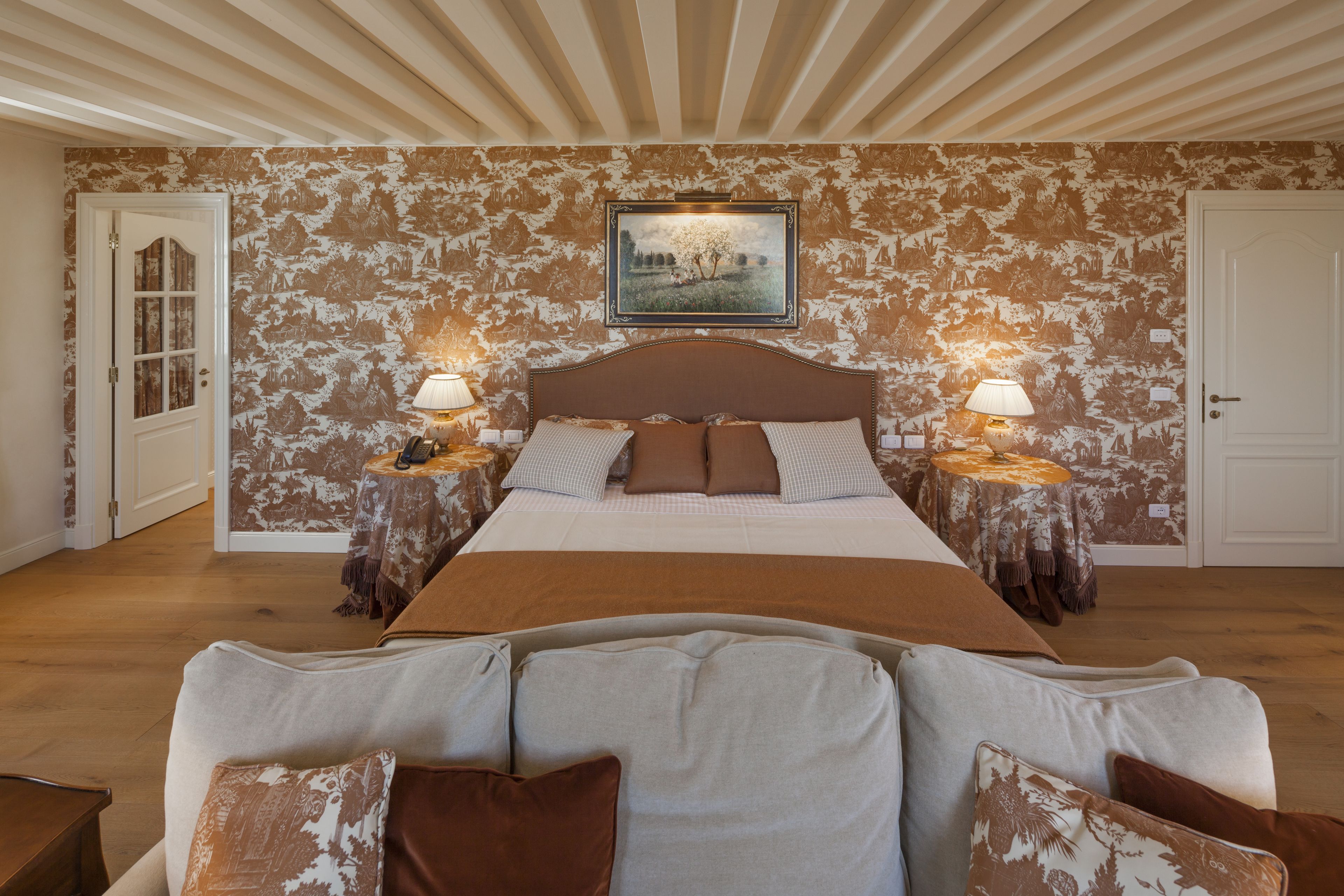 A bedroom with a large bed, two bedside tables with lamps, a framed picture on the wall, and a patterned wallpaper.