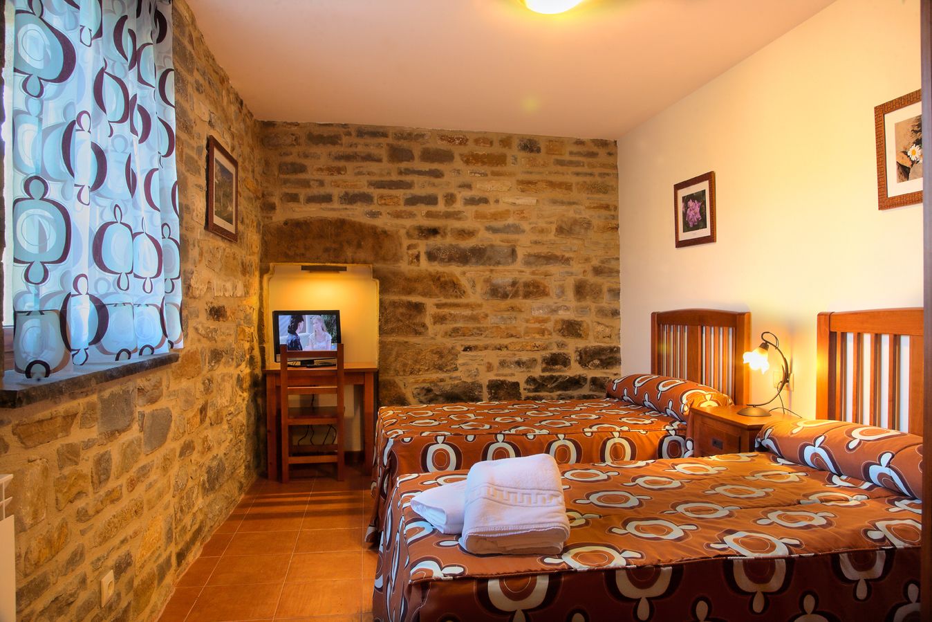Une pièce avec un mur en pierre, un téléviseur posé sur un meuble en bois, deux lits avec une literie à motifs, une table de chevet avec une lampe et des tableaux encadrés accrochés au mur.