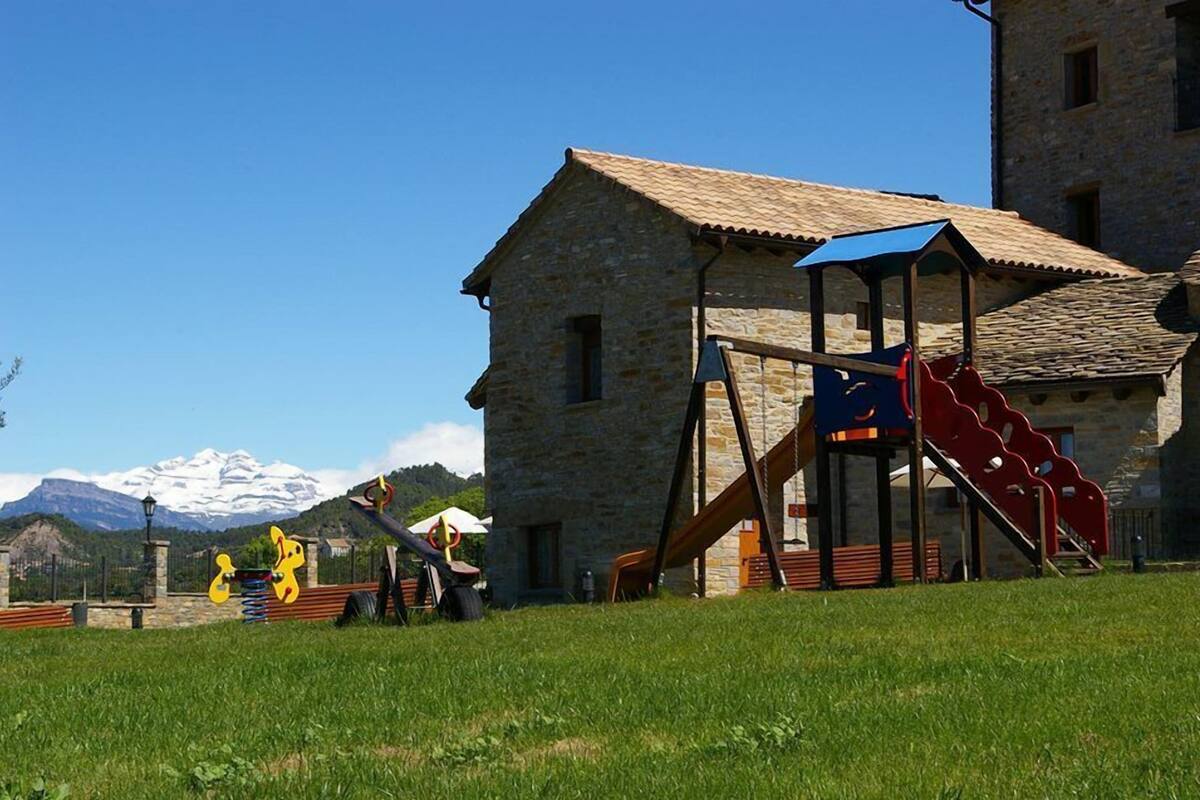 A stone building with a playground featuring a slide and swings, set against a backdrop of mountains and clear sky.