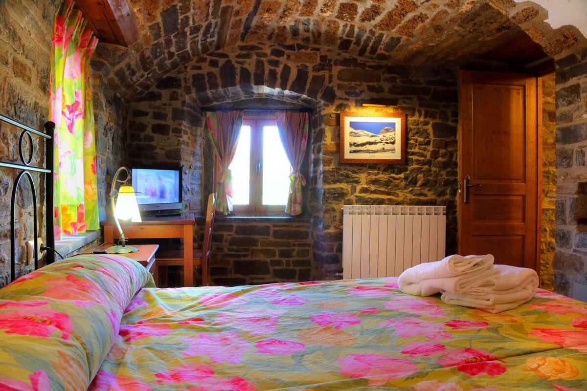 A cozy room with a stone wall, a wooden door, a bed with floral bedding, a desk with a lamp, and a television.