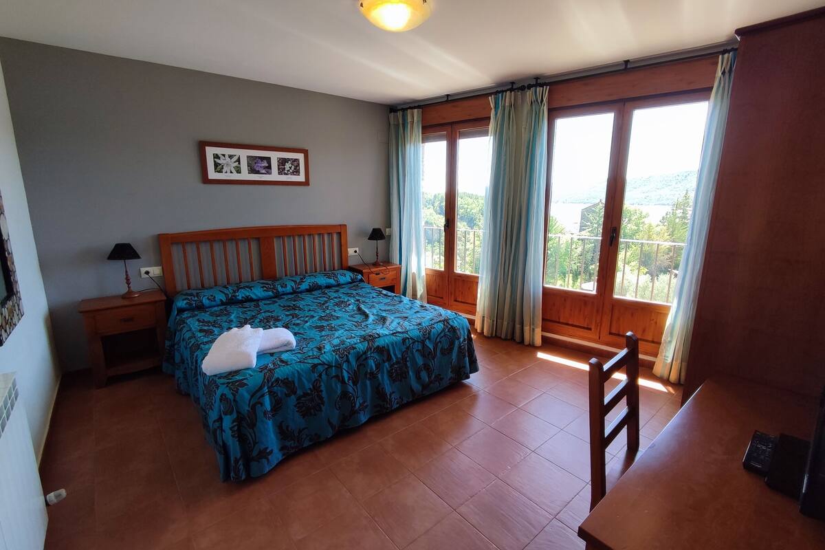 A bedroom with a large bed, a wooden headboard, a nightstand with a lamp, a desk, and a window with curtains.