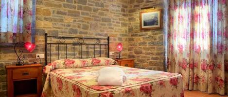 A bedroom with a stone wall, floral bedspread, a bedside table with a lamp, and curtains.
