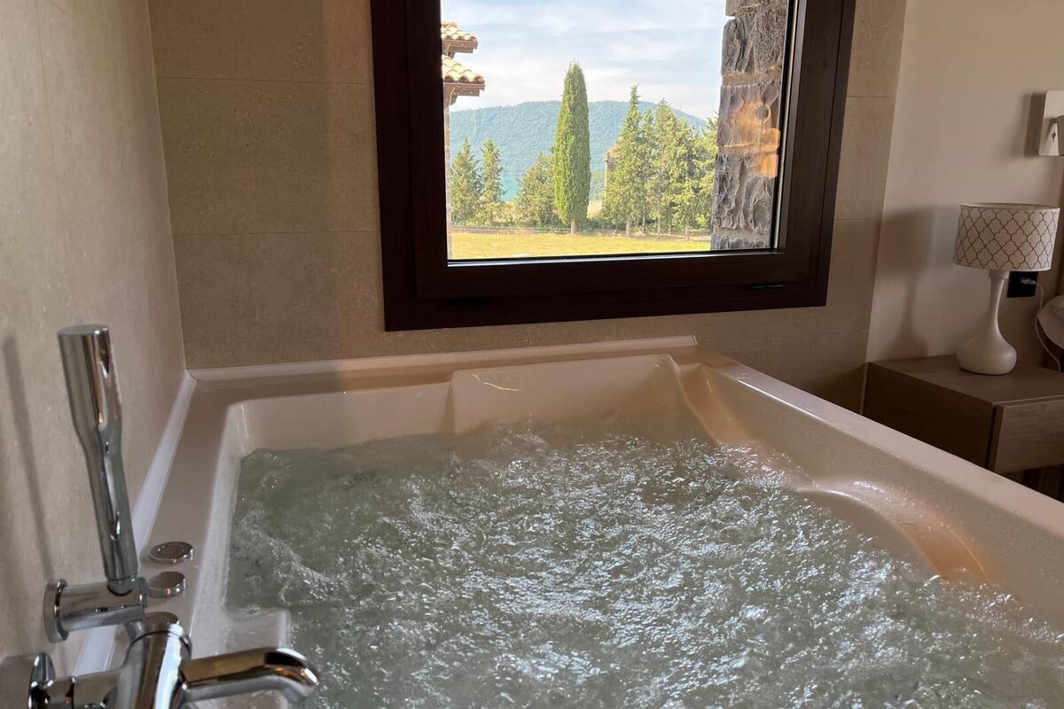 A bathtub with running water, a window with a view of trees and mountains, and a lamp on a bedside table.