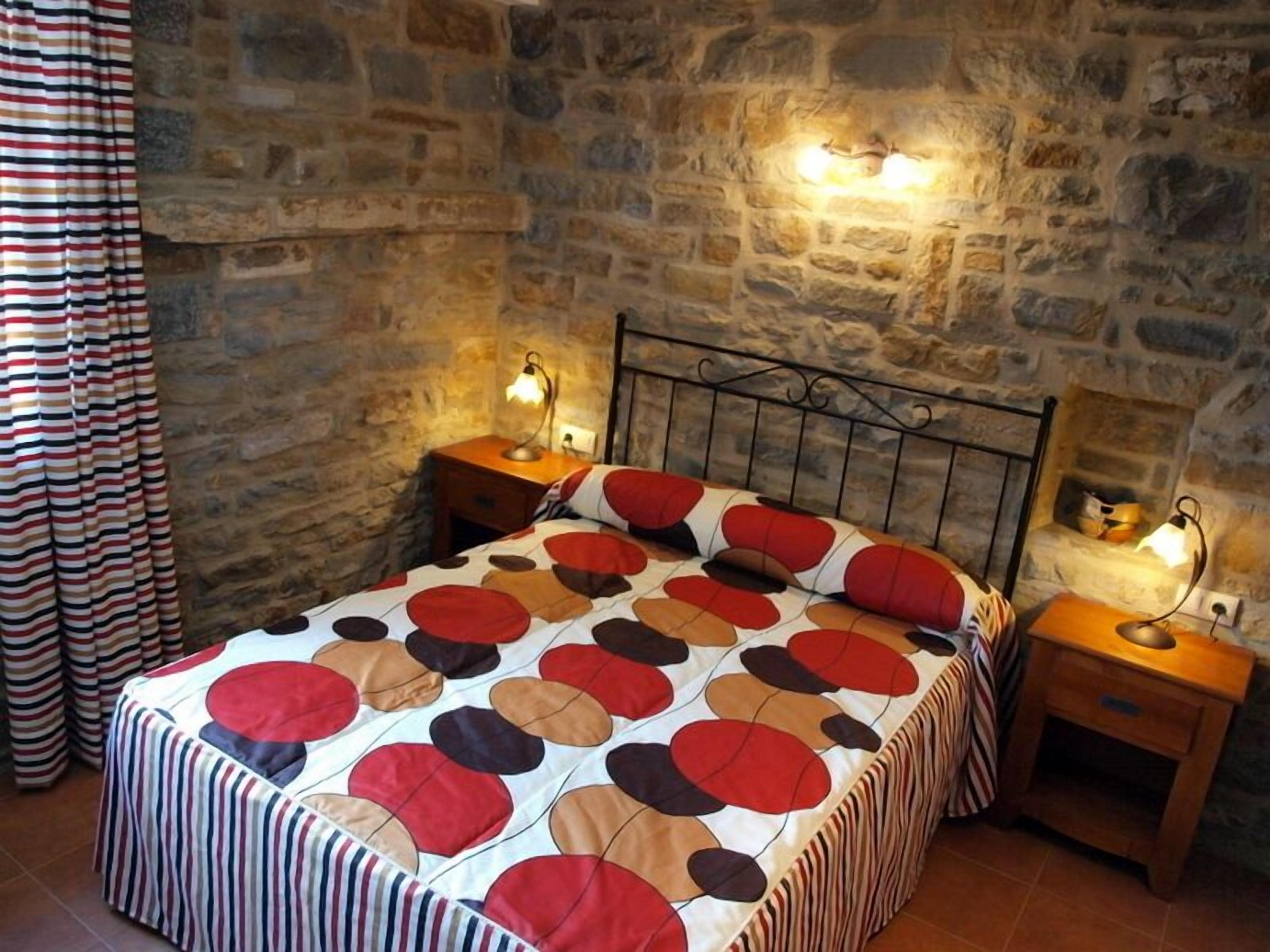 Un dormitorio con pared de piedra, una cama con colcha estampada, dos mesitas de noche con lámparas y una cortina a rayas.